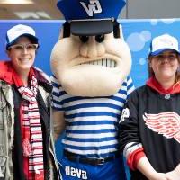Two alumnae pose, wearing the Red Wings GVSU hat with Louie the Laker at the Detroit Red Wings GVSU Night